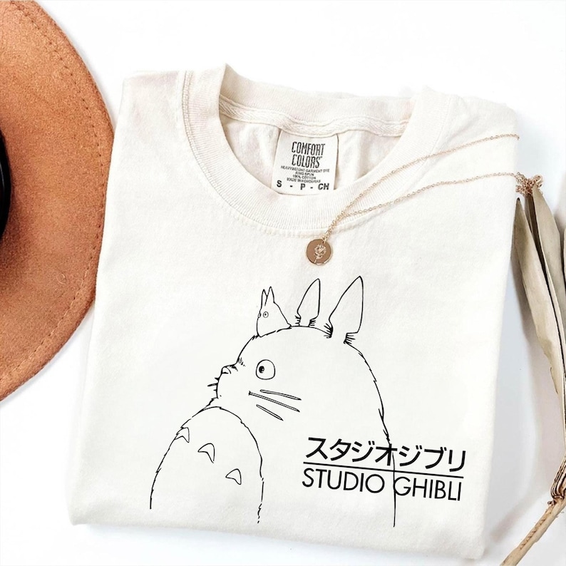 May include: Cream-colored t-shirt featuring a black line drawing of a cartoon character with the Japanese text "スタジオジブリ STUDIO GHIBLI". A gold necklace with a pendant is draped across the shirt. The shirt is folded, with a brown hat visible in the background.