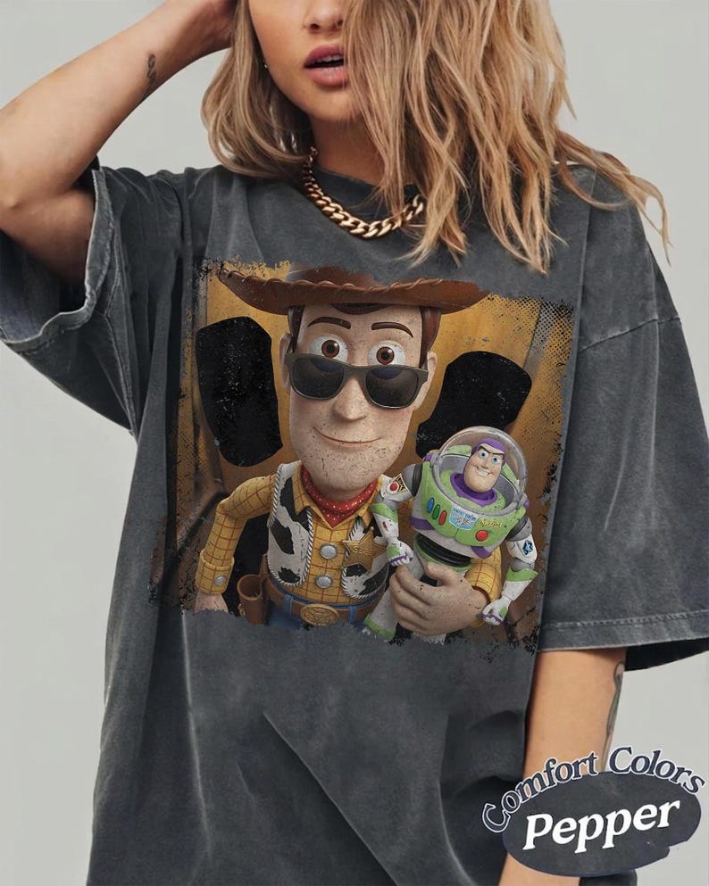 Op de afbeelding: Een antracietgrijs, acid-washed t-shirt met een grafische print van Woody die een zonnebril draagt en Buzz Lightyear vasthoudt. Het t-shirt heeft een ronde hals en korte mouwen. De tekst Comfort Colors Pepper staat rechtsonder op het shirt.