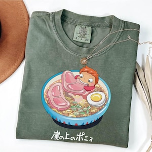 Puede incluir: Camiseta Comfort Colors verde oliva con una ilustraci&oacute;n de dibujos animados de un personaje en un cuenco de ramen con carne, fideos y un huevo. El texto japon&eacute;s "崖の上のポニョ" est&aacute; debajo. Un collar dorado est&aacute; sobre la camiseta.