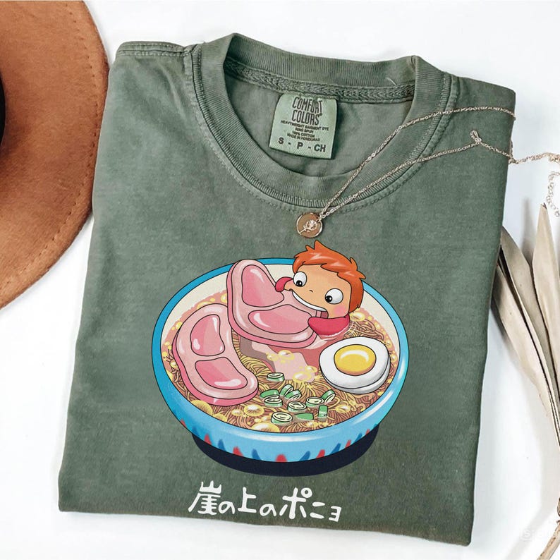 Puede incluir: Camiseta Comfort Colors verde oliva con una ilustraci&oacute;n de dibujos animados de un personaje en un cuenco de ramen con carne, fideos y un huevo. El texto japon&eacute;s "崖の上のポニョ" est&aacute; debajo. Un collar dorado est&aacute; sobre la camiseta.