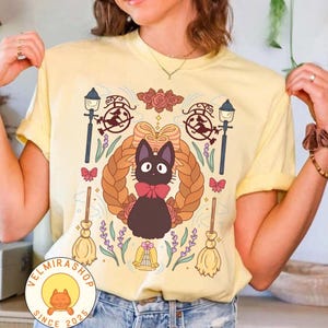 May include: Yellow t-shirt featuring a cartoon cat with a red bow tie, surrounded by a braided wreath, brooms, and floral elements. The design includes a witch on a broom and the text "VELMIRASHOP SINCE 2025".