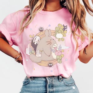 May include: Pink crew neck sweatshirt featuring a cartoon illustration of Totoro and other characters from the animated film My Neighbor Totoro. The design includes a large gray Totoro, smaller characters, flowers, and leaves. The sweatshirt is made of a soft material.