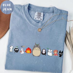 May include: Blue jean colored t-shirt featuring a line of cartoon characters. The shirt has a crew neck and short sleeves. A gold necklace with a pendant is draped over the shirt. The shirt has the Comfort Colors brand tag.