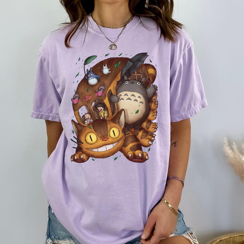 May include: A light purple t-shirt featuring a colorful graphic of characters from a popular animated film. The design includes a large brown creature, a smiling cat, and other animated figures. The shirt is made of soft material.