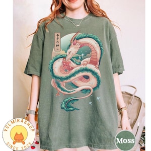 May include: Moss green t-shirt featuring a detailed illustration of a white and red dragon with teal accents, set against a backdrop of a blue sky and a Japanese text. The shirt has a round neck and short sleeves.