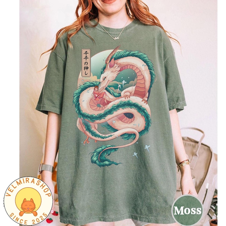 May include: Moss green t-shirt featuring a detailed illustration of a white and red dragon with teal accents, set against a backdrop of a blue sky and a Japanese text. The shirt has a round neck and short sleeves.