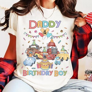 Puede incluir: Una camiseta color crema con un gr&aacute;fico colorido que dice "DADDY OF THE BIRTHDAY BOY". El gr&aacute;fico presenta coches de dibujos animados, una tarta de cumplea&ntilde;os, globos y confeti, creando un tema festivo de cumplea&ntilde;os.