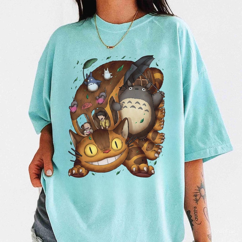 May include: A light blue t-shirt featuring a colorful illustration of characters from the animated film "My Neighbor Totoro." The design includes Totoro, a cat bus, and other forest spirits. The shirt is made of a soft material.