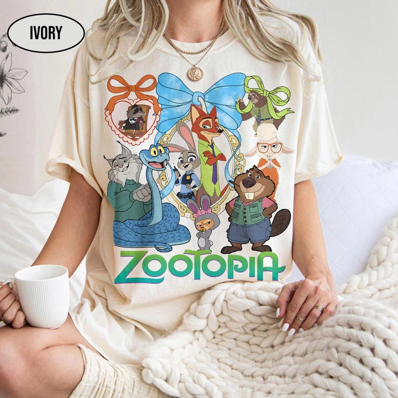 Puede incluir: Camiseta de color marfil con un gr&aacute;fico colorido de personajes de la pel&iacute;cula animada "Zootopia". El dise&ntilde;o incluye un lazo azul, un marco en forma de coraz&oacute;n y la palabra "Zootopia" en verde. La camiseta tiene mangas cortas.