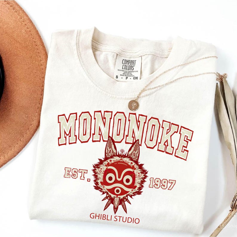 May include: Cream-colored t-shirt featuring the word "MONONOKE" in a collegiate-style font, with a red and cream-colored illustration of a wolf-like creature. The text "EST. 1997" and "GHIBLI STUDIO" are also printed on the shirt. A gold necklace rests on the shirt.