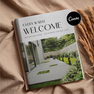 May include: A magazine with the words "A Very Warm Welcome" on the cover, featuring a modern house exterior. The magazine is on a beige surface with dried plants in the background. The Canva logo is in the upper right corner.