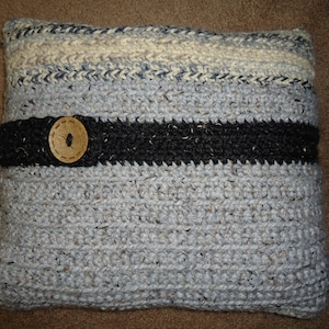 Op de afbeelding: Een rechthoekig, handgemaakt gehaakt kussen in tinten grijs, blauw en crème. Een dikke zwarte band met een houten knoopdetail accentueert het midden. Het kussen is een decoratief woonaccessoire.