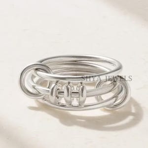 May include: A silver ring with a unique design. The ring features multiple intertwined bands and decorative elements. The ring is made of a shiny, metallic material and is displayed on a light-colored surface. The ring has the text "SHYA JEWELS" on it.