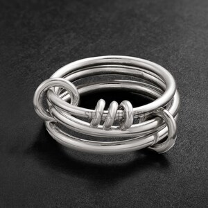 Puede incluir: Un anillo plateado con un diseño único. El anillo presenta múltiples bandas entrelazadas con elementos en bucle y enrollados. El metal pulido refleja la luz, creando una apariencia brillante y llamativa. El anillo está sobre un fondo oscuro.