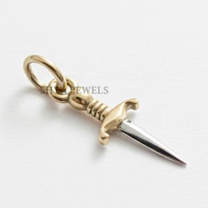May include: A gold-coloured dagger pendant with a silver blade. The pendant has a small loop for attaching to a chain. The word "JEWELS" is visible on the pendant. The pendant is likely made of metal and is designed to be worn as jewellery.