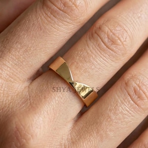 Peut inclure: Une bague dorée avec un design unique en forme de nœud papillon. La bague est portée sur un doigt, mettant en valeur sa surface polie et réfléchissante. Le design présente une indentation centrale, créant une forme distinctive. La bague est un bijou.