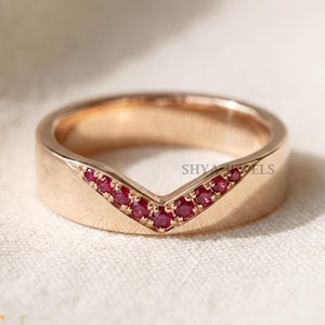 May include: A rose gold ring with a V-shaped design. The ring features a row of small, vibrant red gemstones set into the V. The band is smooth and polished, with a modern, minimalist aesthetic. The ring is likely a piece of jewellery.