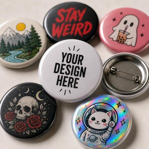 May include: A collection of six circular pin-back buttons featuring various designs: a mountain landscape, a black button with red text reading STAY WEIRD, a pink ghost with boba tea, a white button with black text YOUR DESIGN HERE, a skull with roses, and a holographic cat astronaut.