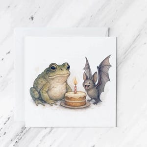 Op de afbeelding: Een vierkante wenskaart met een aquarelillustratie van een groene pad die naast een kleine grijze vleermuis zit. Tussen hen in staat een ronde beige verjaardagstaart op een bordje met een enkele rood-wit gestreepte kaars.