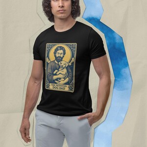 Op de afbeelding: Zwart t-shirt met een afbeelding van een man die een gele Labrador puppy vasthoudt, met de tekst "THE DEVOTED DOG DAD". De afbeelding is omlijst in een goud en blauw ontwerp. Het shirt is gemaakt van een zacht materiaal.