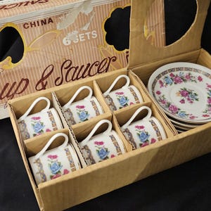 May include: A boxed set of six fine china cups and saucers. The cups and saucers are white with a floral design featuring blue and pink roses. The box is beige with the words "Cup & Saucer" printed on it.