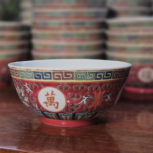 May include: A decorative ceramic bowl with a red and white design. The bowl has a white interior, a red exterior with a repeating pattern, and a Chinese character in a white circle. Other bowls are stacked in the background.