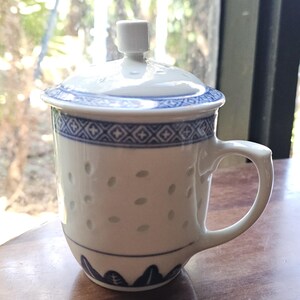 May include: White ceramic tea cup with a matching lid. The cup has a blue and white floral design around the rim and base, with small dotted perforations. The handle is curved, and the lid has a small knob.