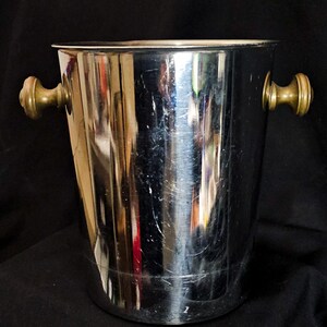 May include: A shiny, stainless steel ice bucket with brass handles. The bucket has a cylindrical shape and is designed to hold ice for chilling beverages. The handles are small and round, adding a touch of elegance to the design.