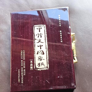 Może przedstawiać: Prostokątne, ciemnobordowe pudełko ze złotymi okuciami. Pudełko zawiera chińskie znaki i angielski tekst z napisem "CHINESE CHESS WITH ORACLE BONE INSCRIPTIONS". Pudełko jest prawdopodobnie przeznaczone na zestaw szachów.