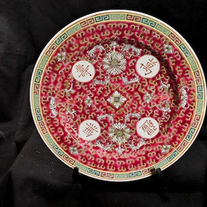 May include: A decorative red and white porcelain plate with intricate floral and geometric patterns. The plate features four white medallions with Chinese characters and a detailed border. The plate is displayed on a black stand.