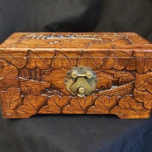 May include: A rectangular wooden box with detailed carvings. The box is a warm brown colour and features relief carvings of landscapes and leaf patterns. A brass clasp secures the lid. The box is likely used for storing small items.