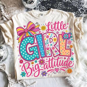 May include: A cream-colored t-shirt featuring a colorful graphic with the text Little Girl Big attitude. The design includes a large striped bow, polka-dot lettering in teal and orange, and various floral accents in pink, yellow, and purple.