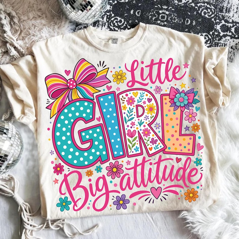 May include: A cream-colored t-shirt featuring a colorful graphic with the text Little Girl Big attitude. The design includes a large striped bow, polka-dot lettering in teal and orange, and various floral accents in pink, yellow, and purple.