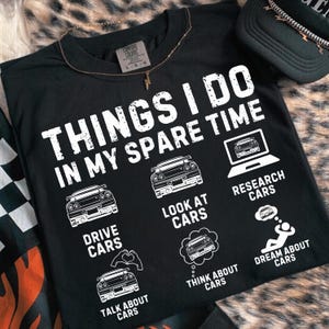 May include: Black t-shirt featuring the phrase "THINGS I DO IN MY SPARE TIME" with car-themed illustrations. The shirt includes graphics for "DRIVE CARS", "LOOK AT CARS", "RESEARCH CARS", "TALK ABOUT CARS", "THINK ABOUT CARS", and "DREAM ABOUT CARS".