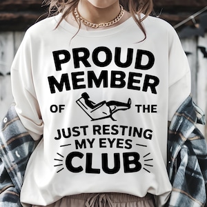 Op de afbeelding: Crèmekleurige sweatshirt met de tekst "PROUD MEMBER OF THE JUST RESTING MY EYES CLUB" in vette zwarte letters. De afbeelding toont een persoon die in een stoel ligt. Een gouden ketting is zichtbaar.