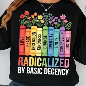 Op de afbeelding: Zwart sweatshirt met een afbeelding van kleurrijke boeken, elk met een woord: Liefde, Vrede, Inclusie, Vriendelijkheid, Verandering, Diversiteit, Eenheid, Compassie en Rechtvaardigheid. Bloemen groeien uit de boeken. De tekst luidt "GERADICALISEERD DOOR BASISDECENTIE".