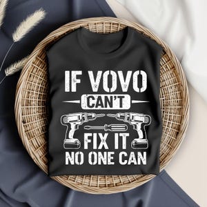 Pode incluir: Camiseta preta com o texto branco "IF VOVO CAN'T FIX IT NO ONE CAN". O design inclui duas furadeiras sem fio e uma chave de fenda. A camiseta é exibida em uma cesta de vime.