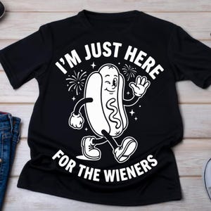 May include: A black short-sleeved t-shirt featuring a white graphic of a cartoon hot dog character waving and winking. The text "I'M JUST HERE FOR THE WIENERS" is printed in a bold, arched font above and below the central hot dog illustration.