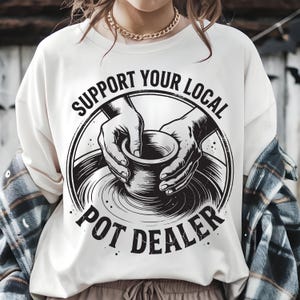 May include: Cream-colored sweatshirt with a black graphic that reads "SUPPORT YOUR LOCAL POT DEALER." The graphic features hands shaping a pot on a pottery wheel. The sweatshirt is worn by a person with a gold chain necklace and a plaid shirt.