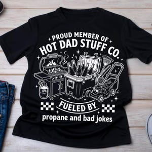May include: Black t-shirt with a white graphic that reads "PROUD MEMBER OF HOT DAD STUFF CO." and "FUELED BY propane and bad jokes." The graphic includes a grill, cooler with bottles, lawnmower, and rake.