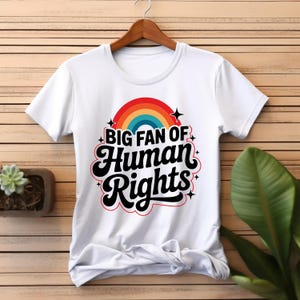 Puede incluir: Camiseta blanca con el texto "BIG FAN OF Human Rights" en fuente retro, con un arcoíris. La camiseta está colgada en una percha de madera sobre un fondo de madera.