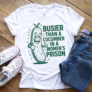 Puede incluir: Camiseta blanca con una ilustración de pepino verde y el texto "BUSIER THAN A CUCUMBER IN A WOMEN'S PRISON." La imagen también muestra vaqueros azules y zapatillas blancas.