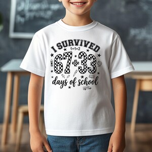 以下が含まれることがあります： 「I SURVIVED 1+1=2 67+33 days of school」の黒い文字が入った白いTシャツ。デザインには星、クレヨン、鉛筆、地球儀、スクールバスが含まれています。半袖のシャツです。