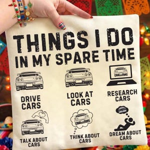 May include: Cream-coloured square cushion with the text "THINGS I DO IN MY SPARE TIME" and car illustrations. The cushion features the phrases "DRIVE CARS", "LOOK AT CARS", "RESEARCH CARS", "TALK ABOUT CARS", "THINK ABOUT CARS", and "DREAM ABOUT CARS".
