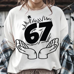 May include: Cream-colored sweatshirt with a black graphic of the number 67, stylized with a star and lines, and two hands cupping the number. The sweatshirt has long sleeves and a relaxed fit.