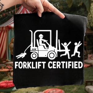 Könnte beinhalten: Schwarzer Stoff mit einer weißen Grafik eines Gabelstaplers, einer Person, die ihn bedient, und zwei laufenden Figuren. Der Text "FORKLIFT CERTIFIED" befindet sich darunter. Der Stoff wird von einer Hand gehalten.