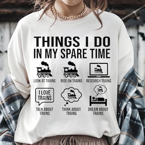 May include: Cream-coloured sweatshirt featuring the text "THINGS I DO IN MY SPARE TIME" and train-related illustrations. The design includes train icons, a laptop, and speech bubbles with phrases like "TALK ABOUT TRAINS".