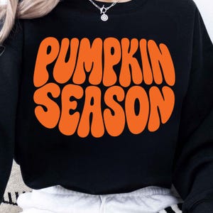 Może przedstawiać: Czarna bluza z długim rękawem z napisem PUMPKIN SEASON wydrukowanym dużymi, pogrubionymi, pomarańczowymi literami na klatce piersiowej. Czcionka ma falisty, zaokrąglony styl. Odzież posiada okrągły dekolt oraz długie rękawy z prążkowanymi mankietami.