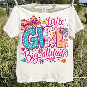 May include: A cream-colored short-sleeve t-shirt featuring a colorful graphic print. The design includes the text "Little GIRL Big attitude" in pink and blue, decorated with polka dots, floral patterns, a large striped bow, and small flowers.