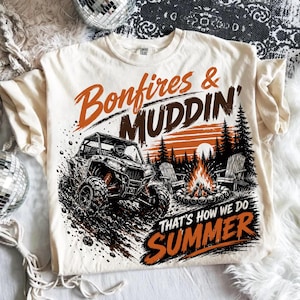 May include: A cream-coloured t-shirt featuring a graphic print of an off-road vehicle in mud, a campfire with two chairs, and pine trees. The text reads Bonfires & MUDDIN' THAT'S HOW WE DO SUMMER in orange and brown lettering across the front.